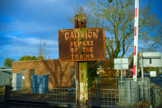 Vorsichtsschild an einem Bahnübergang-Zaun neben Bäumen, Pfählen, einem Gebäude mit Fenstern, Containern, einem Straßenpfahl, Fahrzeugen auf der Straße und einem bewölkten Himmel.