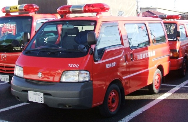 Drei rote Rettungswagen mit weißen und schwarzen Markierungen nebeneinander auf einem Parkplatz geparkt, mit Gebäuden und Polen im Hintergrund unter einem klaren blauen Himmel.