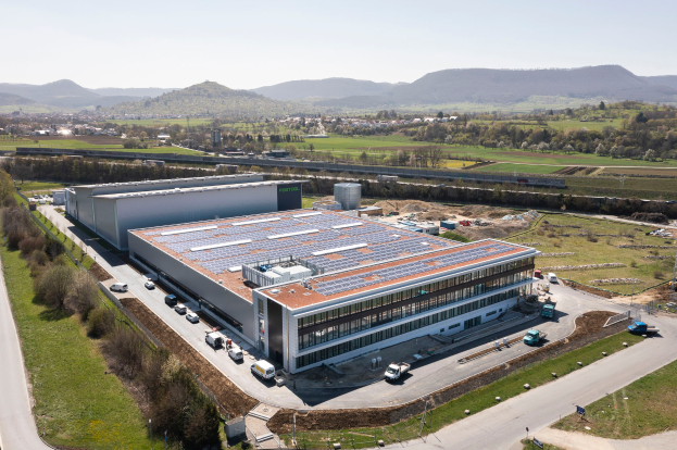Eine Luftaufnahme eines großen Fabrikgebäudes mit Solarpaneelen auf dem Dach, umgeben von Fahrzeugen, Bäumen und anderen Gegenständen, mit Hügeln und einem klaren blauen Himmel im Hintergrund.