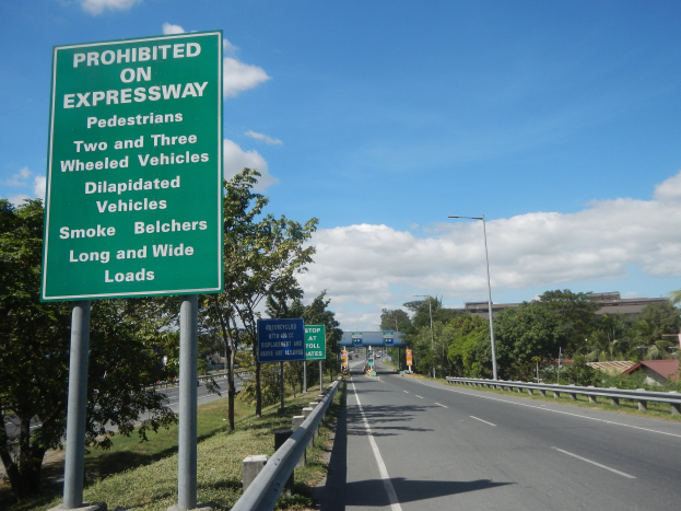 Grünes Highway-Schild mit der Aufschrift "Verboten auf der Autobahn" mit Fahrzeugen, Laternenmästen, Bäumen, Gebäuden und bewölktem Himmel im Hintergrund.