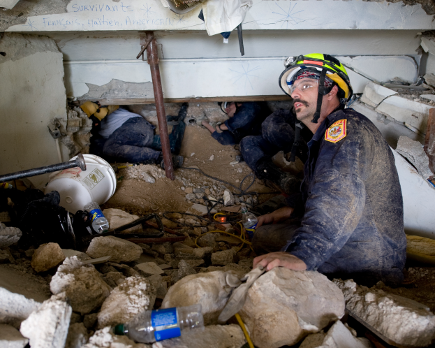 Ein Feuerwehrmann in einem Helm sitzt zwischen Trümmern, darunter Steine und Flaschen, mit einer Wand im Hintergrund, auf der ein Text zu sehen ist.