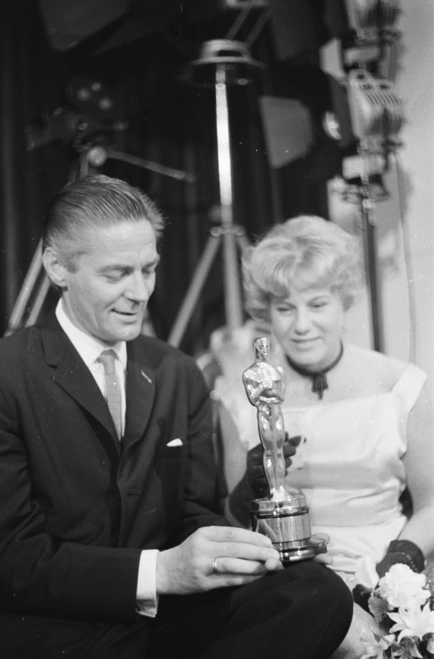 Ein Schwarz-Wei├č-Bild eines formell gekleideten Mannes, der eine Oscar-Statue hölt und eine Frau, die einen Blumenstrau├č hölt, nebeneinander sitzen mit beleuchtetem Hintergrund.