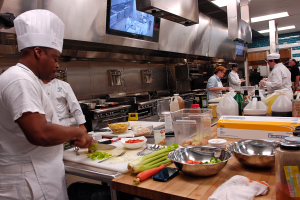 Eine Gruppe von Köchen in einer Küche bei der Zubereitung von Speisen auf einem Schneidbrett, wobei ein Koch im Vordergrund in einer weißen Schürze und Mütze mit einem Messer in der Hand zu sehen ist und von verschiedenen Utensilien, Schüsseln, Gemüse, einem Handy, Flaschen und anderen Gegenständen umgeben ist, mit einem Fernseher und Deckenleuchten im Hintergrund.