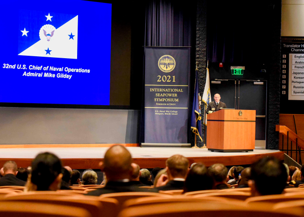 Admiral Mike Gilday steht an einem Podium mit einem Mikrofon und einer Tafel mit Text und Logo, spricht vor einer sitzenden Menge bei der Internationalen Seemacht-Symposium 2021, mit Flaggen, einem Bildschirm mit Text und einer Treppe mit Geländern im Hintergrund.