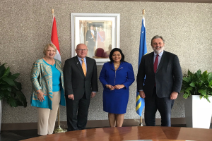 Eine Gruppe strahlender Menschen um einen Tisch mit Papieren herumstehend, umgeben von Fahnen, Topfpflanzen und einem Foto auf dem Wandregal, bei der Unterzeichnung eines Memorandums von Verständigung zwischen den Vereinigten Staaten und Kanada.