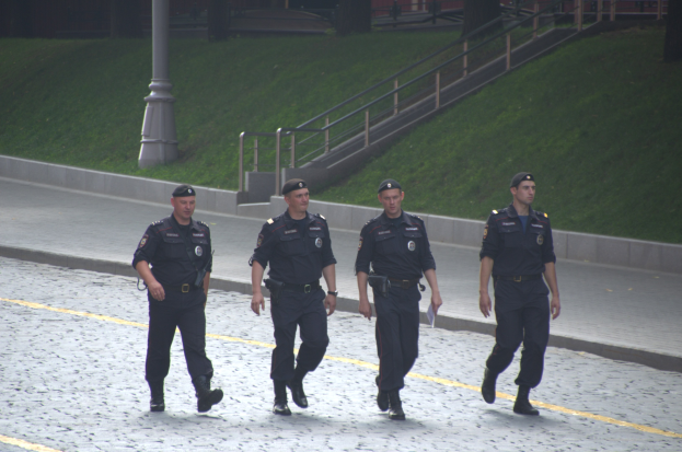 Vier Polizisten in Uniform gehen eine Straße entlang, halten Papiere und haben eine Stange, Treppe, Geländer, Gras, Bäume und ein Gebäude im Hintergrund.