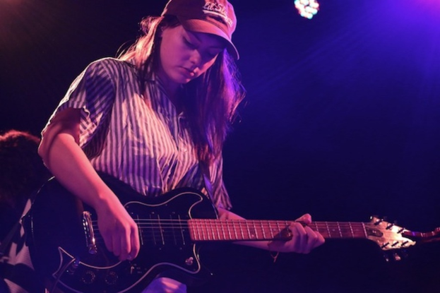 Eine Frau mit einer MÃ¼tze spielt eine Gitarre auf der BÃ¼hne bei einem Musikkonzert, mit zahlreichen Lichtern, die den Hintergrund erhellen.