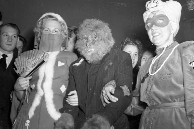 Eine Gruppe von Menschen, die zusammen auf einer Party stehen, alle tragen Masken und einige halten Fächer in der Hand, dargestellt in Schwarz-Weiß.