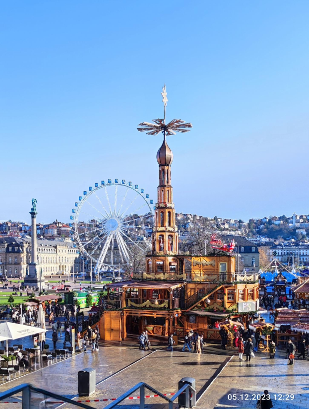 Ein lebhafter Stadtplatz mit einem zentralen Riesenrad, umgeben von Gebäuden, Bäumen und einem klaren blauen Himmel, mit zahlreichen Menschen, Zelten und Geländern und einem Wasserzeichen, das darauf hinweist, dass es eines der besten Weihnachtsmärkte in Europa ist.
