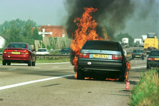 Ein Auto von Flammen umgeben auf der Straße, mit anderen Fahrzeugen in der Nähe, Bäumen, Gebäuden und einem klaren blauen Himmel im Hintergrund und einem Feuerlöscher auf der rechten Seite.