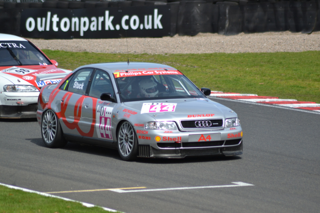 Zwei Autos auf einer grasumsäumten Rennstrecke mit Helmträgern im Hintergrund, umgeben von Reifen und einer Texttafel.