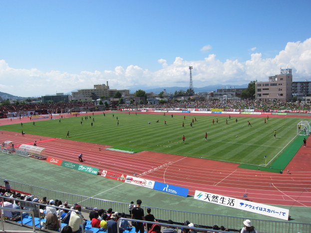 Großes Stadion voller Zuschauer bei einem Fußballspiel mit sitzenden und stehenden Zuschauern, Geländern, Beschriftungen und umgeben von Gebäuden, Bäumen, Türmen, Hügeln und einem bewölkten Himmel.