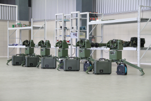 Eine Gruppe von Militärrobotern ist auf einem Boden positioniert, umgeben von Koffern und anderen Gegenständen auf Regalen, mit einer Wand und einem Geländer im Hintergrund.