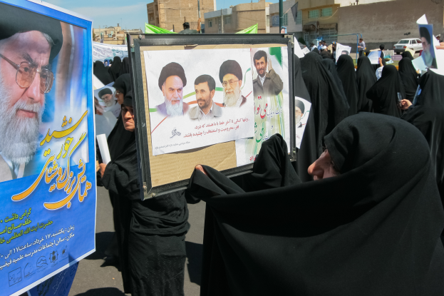 Eine Gruppe von Frauen in schwarzen Abayas hält Plakate mit Bildern von Ayatollah Ali Khamenei hoch, im Hintergrund sind Gebäude, Pfosten, Fahrzeuge und ein klarer blauer Himmel zu sehen.