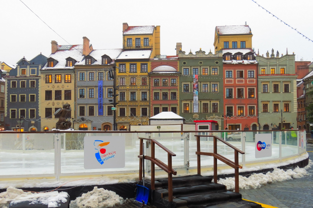 Eisbahn in einem Stadtplatz umgeben von Gebäuden, Treppen, Geländern, Informationsschildern, schneebedeckter Fläche, Laternen, Bäumen und einem Himmel als Hintergrund.