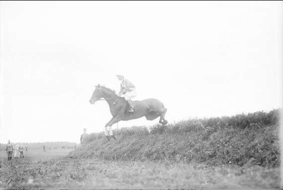 Schwarz-weiß-Foto eines Pferdes und Reiters, die über einen grünen Hügel springen, mit dem Pferd im Galopp und dem Reiter in einem Helm und einem langen Mantel.