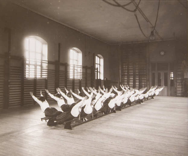 Gruppe von Menschen, die Pilates-Übungen in einem Fitnessstudio mit einer Uhr an der Wand und Fenstern im Hintergrund durchführen.