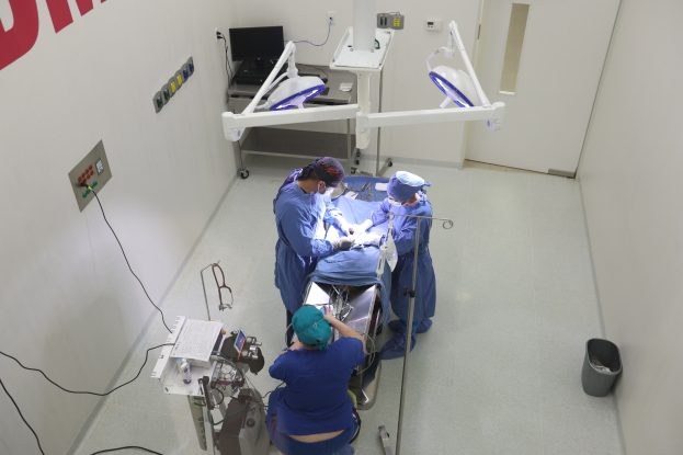 Eine Gruppe von Chirurgen führt einen Eingriff in einem Operationssaal durch, mit einem Patienten auf einem Operationstisch umgeben von medizinischer Ausrüstung, Text an der linken Wand, einem Objekt auf dem rechten Boden und verschiedenen medizinischen Geräten im Hintergrund.