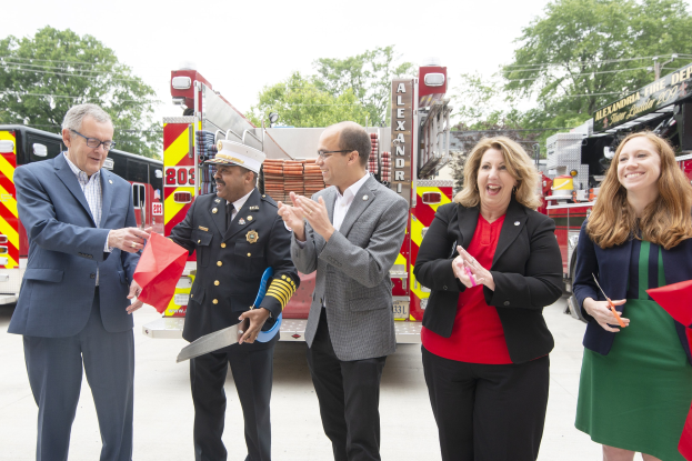 Gruppe von Menschen, die bei einer Einweihungszeremonie der Alexandria Fire Department klatschen und lächeln, mit zwei Personen, die eine Schere und ein rotes Tuch halten, vor einem Feuerwehrauto.