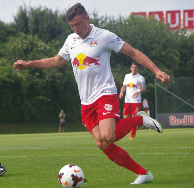 Ein professioneller Fußballspieler schießt einen Ball auf einem Rasenfeld mit Bäumen und einem klaren blauen Himmel im Hintergrund, in der Nähe einer Tafel mit der Aufschrift "RB Leipzig."