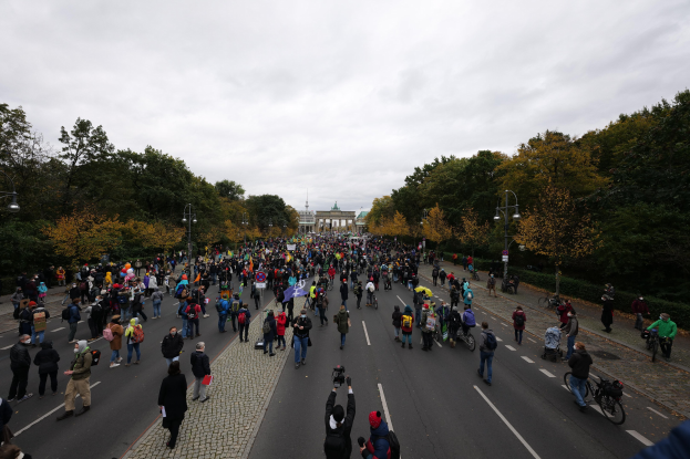 Eine große Gruppe von Menschen marschiert auf einer von Bäumen gesäumten Straße in Berlin, mit Kameras in der Hand und einem Gebäude und einem klaren Himmel im Hintergrund.