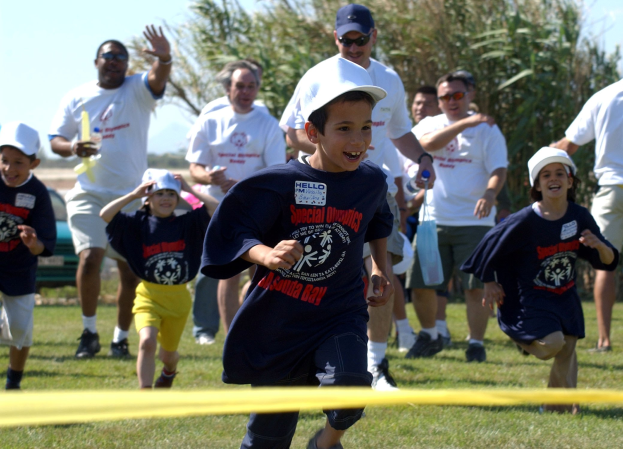 Kinder laufen bei einem Rennen auf einem Rasenfeld mit einem gelben Band im Vordergrund, einige tragen Kappen und Schutzbrillen, andere halten Gegenstände, unter einem klaren blauen Himmel mit Bäumen im Hintergrund.