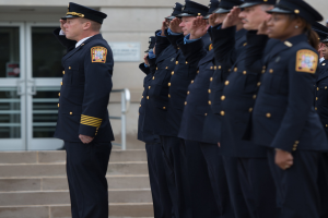 Eine Gruppe von Polizisten in Uniform und Mützen, die in Formation stehen und vor einem Gebäude mit Glastüren und Treppen salutieren.