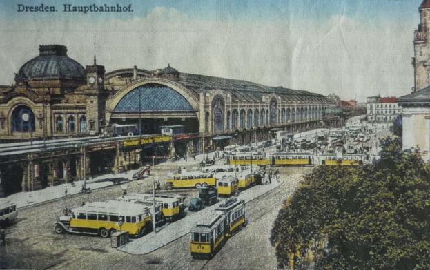 Eine alte Postkarte, die einen belebten Dresdner Bahnhof mit Fahrzeugen, Menschen, Bäumen, Gebäuden und einer bewölkten Himmel zeigt, mit der Aufschrift "Dresden Hauptbahnhof" oben.