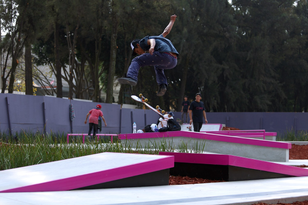 Ein Skateboarder führt einen Sprung auf einer Rampe aus, während eine Gruppe von Zuschauern zusieht, mit verstreuten Gegenständen und einem Hintergrund aus Bäumen, Gebäuden und einem klaren Himmel.
