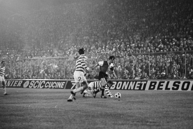 Eine Gruppe von Männern, die auf einem Feld Fußball spielen, mit Zuschauern auf den Rängen und einer Fahne mit der Aufschrift "Celtic v West Bromwich Albion - FA Cup Third Round" in Schwarz-Weiß.