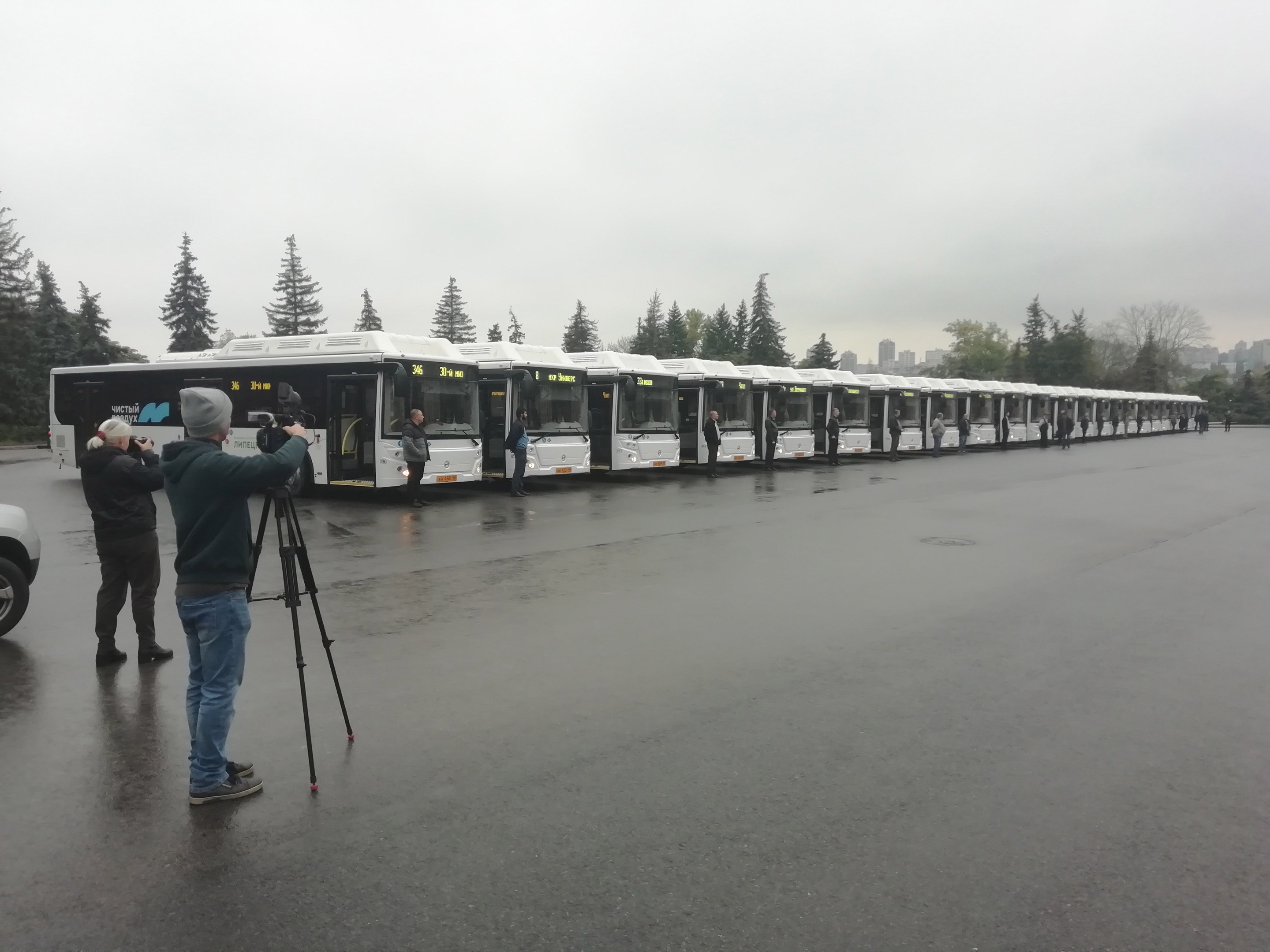 Eine Gruppe von Menschen, die Fotos von einer Reihe von Bussen machen, die auf einer Straße mit Bäumen, Gebäuden und einem klaren blauen Himmel im Hintergrund geparkt sind.