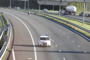 Auto auf der Autobahn mit einem stationären Polizeiwagen auf dem Seitenstreifen, andere Fahrzeuge sichtbar, umgeben von Leitplanken, Strommasten, Schildern und Bäumen.