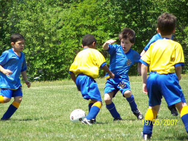 Kinder beim Fußballspielen auf einer grünen Wiese mit Bäumen im Hintergrund und einem Datum in der unteren rechten Ecke.