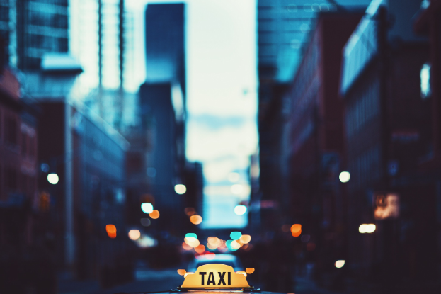Gelbes Taxi fährt nachts auf einer Stadtstraße, mit Gebäuden auf beiden Seiten und einem unscharfen Hintergrund.