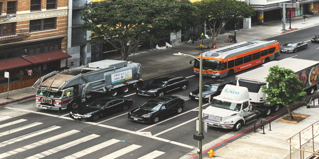 Eine belebte Stadtstraße mit verschiedenen Fahrzeugen, Fußgängern, Gebäuden, Bäumen und Straßenmöbeln, einschließlich Schildern mit Text.