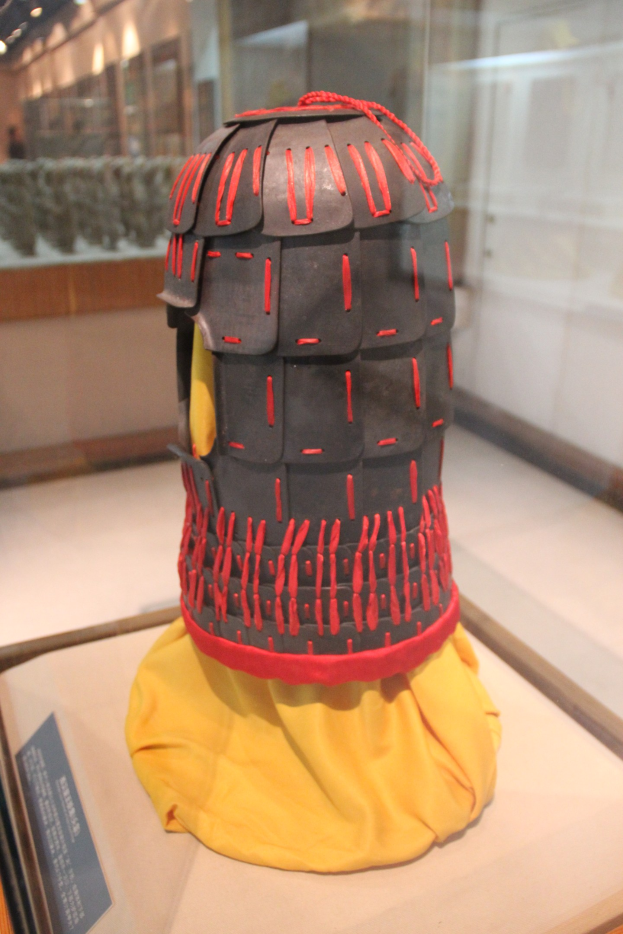 Ein Helm, der in einer Glasvitrine mit einem gelben Tuch darüber ausgestellt ist, begleitet von einer Texttafel links und Hintergrundbeleuchtung und -gegenständen.