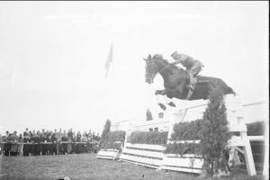 Schwarz-weißes Foto eines Pferdes und Reiters, die über ein Hindernis springen, bei den Royal Ascot Horse Trials im Jahr 1953, mit Zuschauern und einer Fahne im Hintergrund.