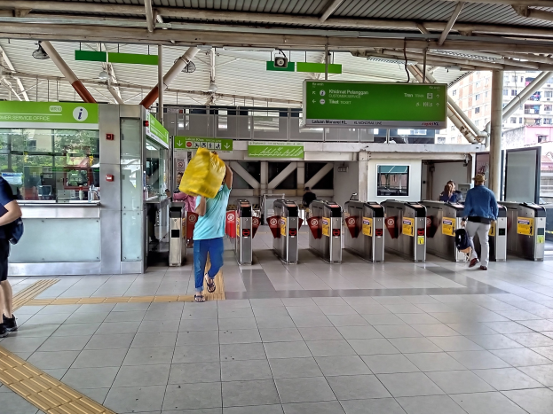 Menschen gehen durch einen Bahnhof mit Metalldetektoren, Hinweisschildern, Metallstangen, Deckenleuchten und Gebäuden mit Fenstern im Hintergrund, einige tragen Taschen.
