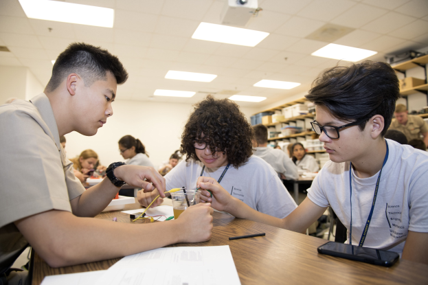 Three students at a table in a classroom work on a project, surrounded by papers, pens, a mobile phone, and other items, with storage racks and ceiling-mounted lights and a projector in the background.