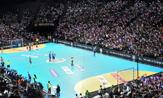 Gruppe von Menschen, die Handball in einem Stadion mit einem Netz auf dem Feld und Zuschauern in den Rängen spielen, während Fotografen das Spiel aufnehmen.