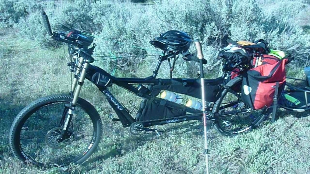Ein Tandemfahrrad mit Gepäck und einem Anhänger, das auf einer grasbewachsenen Fläche positioniert ist.