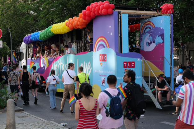 Eine Gruppe von Menschen geht eine Straße entlang neben einem Lastwagen mit bunten Luftballons, mit Schildern an Pfosten entlang der Straße und Bäumen und Gebäuden im Hintergrund, was auf eine Pride-Parade in Paris hindeutet.
