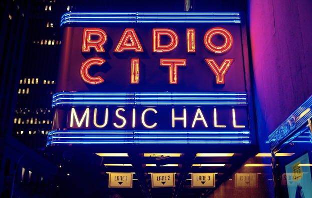 Hell erleuchtetes Radio City Music Hall in New York City mit hohen Gebäuden im Hintergrund, das eine 'Radio City'-Leuchtschrift auf der rechten Seite und mehrere kleinere Schilder darunter zeigt.