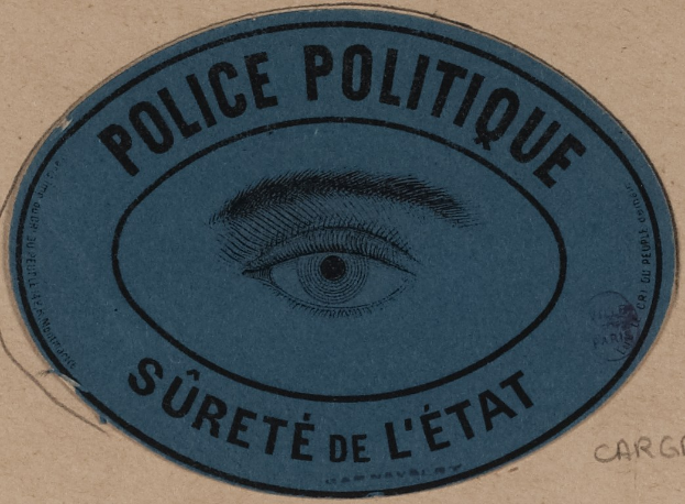 Brauner Karton mit einem blauen Aufkleber, auf dem der Text "Polizei Politique Sûreté de l'État" und ein zentriertes Auge-Symbol steht.