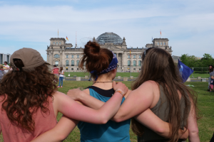 Drei Frauen in verschiedenenfarbigen Kleidern, eine mit einer Mütze, stehen vor dem Reichstaggebäude in Berlin, Deutschland, mit Menschen, Fahnen, Bäumen, Wolken und Himmel im Hintergrund.
