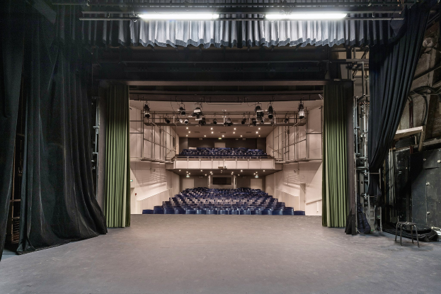 Ein großer Zuschauerraum mit schwarzen Vorhängen, einer Bühne, Deckenlampen und für eine Vorstellung angeordneten Stühlen, mit Gegenständen auf der rechten Seite.