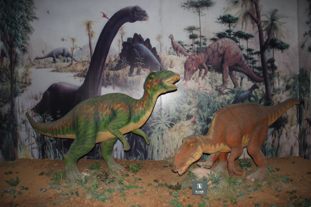 Zwei Dinosaurierskelette in einem Museum ausgestellt, umgeben von Erde und kleinen Pflanzen, mit einer bemalten Wand im Hintergrund mit Bäumen und anderen Dinosauriern.