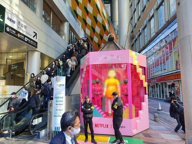 Eine Gruppe von Menschen steht vor einem Gebäude mit Glaswänden, Säulen und Texttafeln, einige tragen Masken, während hinter ihnen eine Rolltreppe mit Menschen nach oben fährt; ein pinker Kasten in der Mitte zeigt Schaufensterpuppen und trägt die Aufschrift "Netflix's new Netflix-Booth in Tokyo."