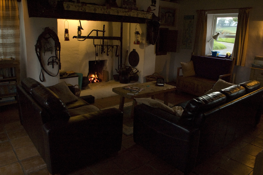 Ein Raum mit einer Couch, einem zentralen Tisch, einem Fenster mit einer Lampe und Vorhang auf der rechten Seite und einer Wand mit Lampen und einem Kamin dahinter.