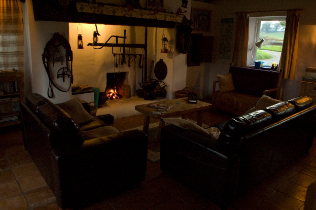 Ein Raum mit einer Couch, einem zentralen Tisch, einem Fenster mit einer Lampe und Vorhang auf der rechten Seite und einer Wand mit Lampen und einem Kamin dahinter.